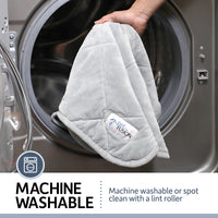 A hand places a PetFusion Microplush Quilted Dog Blanket into a washing machine. Text: MACHINE WASHABLE – easily refresh the ultra-soft blanket by machine washing or spot cleaning with a lint roller, shown with a washing machine icon.