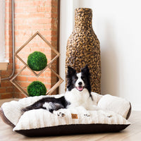 A black and white dog sits happily on a plush white SnooZZy Rustic Luxury Comfy Couch Pet Bed by Snoozzy in a room with a brick wall. A textured vase and geometric wooden sculpture with green spheres enhance the background. The bed is machine washable, blending relaxation with practicality.