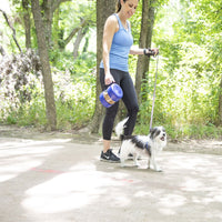 A woman in athletic wear walks her small black and white dog through a forest, holding a blue leash and a Vittles Vault Travel-Trainer Portable Food Storage Container. Sunlight filters through the green trees.