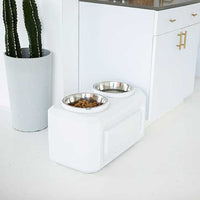 The Vittles Vault Elevated Storage Dog Feeder, featuring Gamma Seal Technology and two stainless steel bowls, is chicly positioned on a white floor beside a white cabinet with gold handles and a cactus in a matching pot, ensuring fresh dog food storage in style.