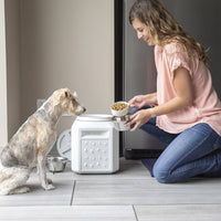 Kneeling on a tiled kitchen floor, a woman in jeans and a pink blouse scoops dog food into a bowl from the Vittles Vault Pawprint Outback Food Storage Container. The BPA-free container is airtight sealed for freshness. Nearby, a small light-colored dog watches eagerly.
