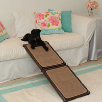 A black dog relaxes on a white sofa with floral pillows. The Gen7Pets Indoor Carpet Mini Ramp by Gen7 offers easy access to the sofa, complementing a cozy room with its blue and beige rug and flowered table.